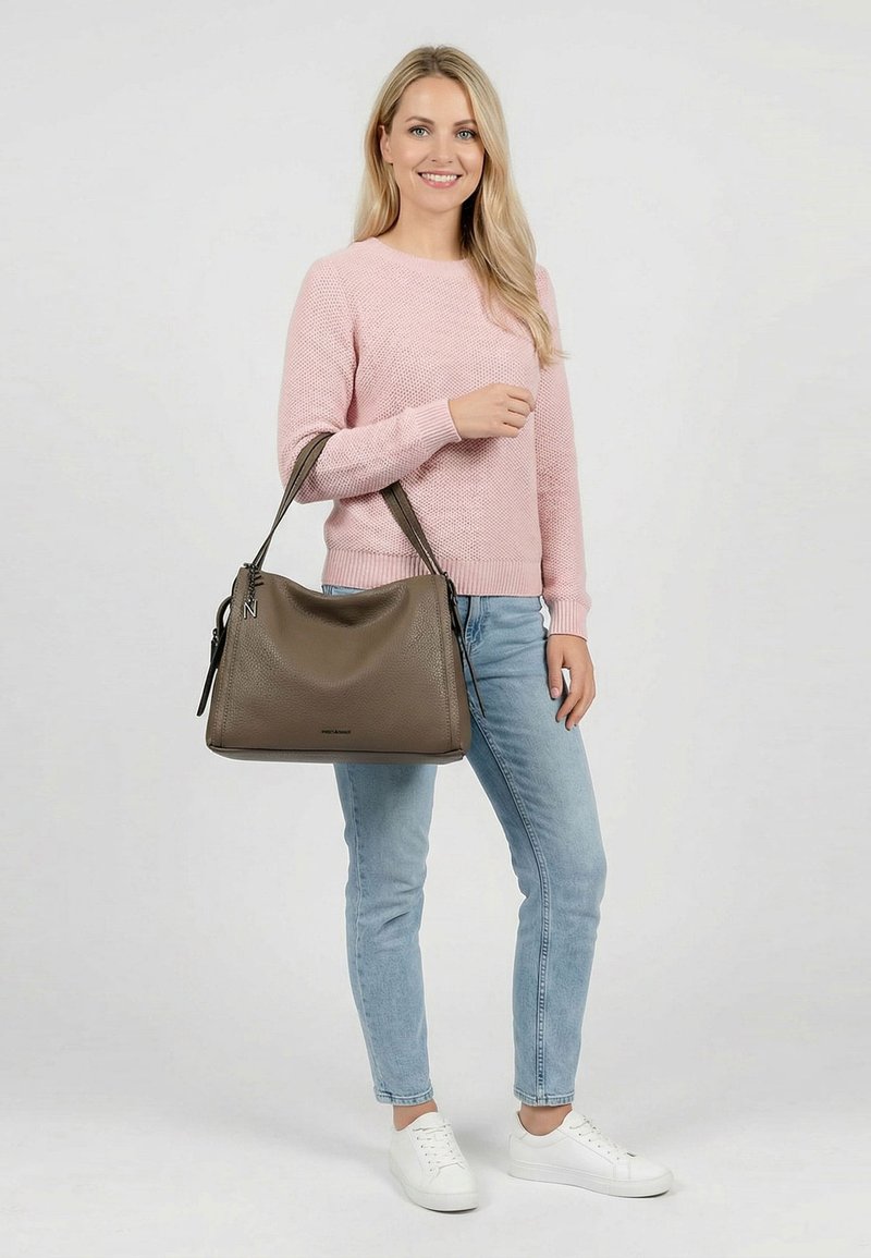 Sieviete rozā džemperī, gaiši zilās džinsās un baltās sporta kurpēs, kas tur lielu brūnu ādas somu uz kreisās rokas un smaida.