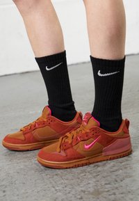 Persona che indossa calzini neri Nike e sneaker marrone-rossastre con vivaci loghi Nike rosa, in piedi su un pavimento di cemento grigio.