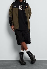 Zielona i czarna polarowa kurtka z zamkiem błyskawicznym na czarnym t-shircie, czarnych spodniach cargo i czarnych botkach za kostkę z teksturowaną podeszwą oraz białymi skarpetkami.