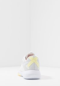 Bílá sportovní bota z prodyšné síťoviny s texturovaným povrchem, zdobená žlutým křížovým vzorem na patě a měkkou podrážkou.