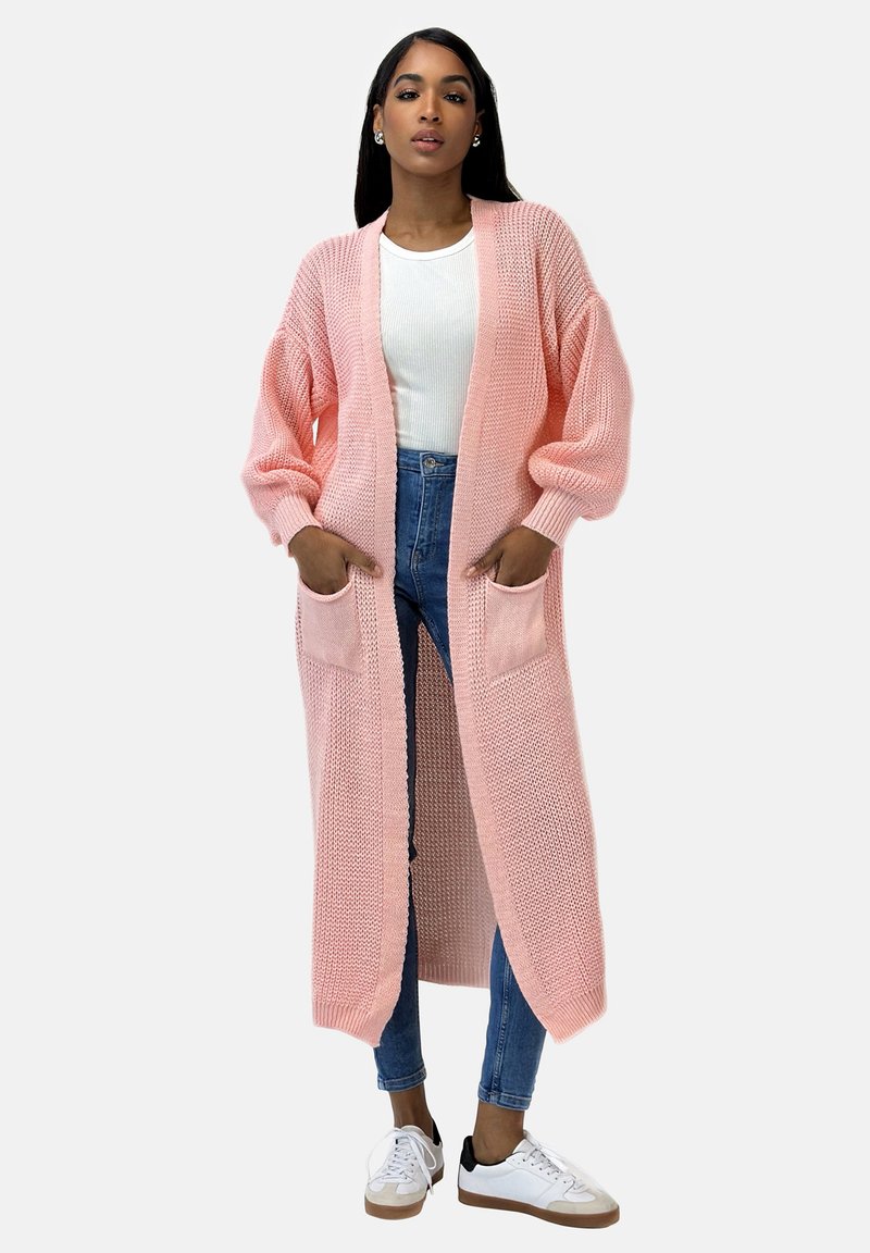 Lång rosa stickad cardigan med öppen framsida, ribbade kanter och två framfickor. Tillsammans med en vit topp och blå jeans, bärs med sneakers.
