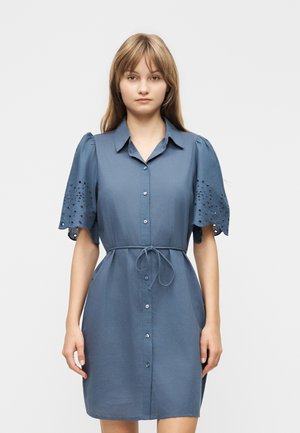 Jeune femme portant une robe bleue boutonnée avec des manches bouffantes en dentelle œillet et une ceinture nouée à la taille, debout devant un fond uni.