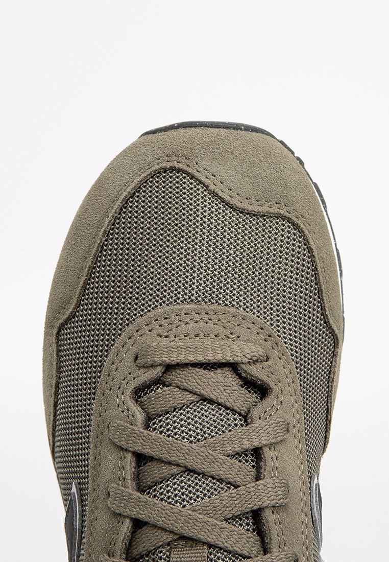 Primo piano della punta di una sneaker taupe con tessuto a rete, sovrapposizioni in suede e lacci incrociati su uno sfondo bianco.