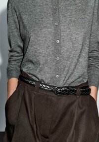 Pull en tricot gris à boutons avec manches longues, associé à un pantalon plié marron et une ceinture en cuir tressé noire.
