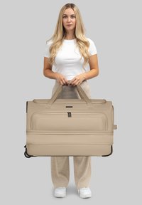 Donna con lunghi capelli biondi, indossando una camicia bianca e pantaloni beige, che tiene in verticale un grande trolley beige per le maniglie.