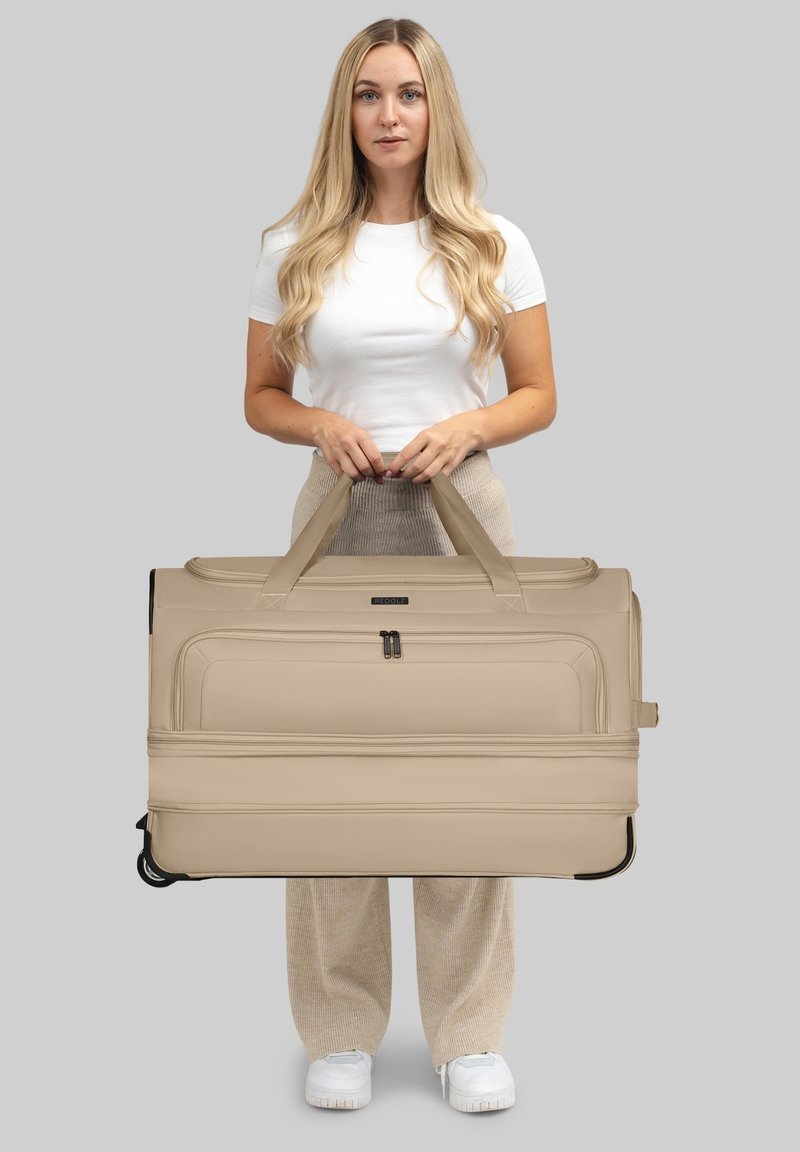Donna con lunghi capelli biondi, indossando una camicia bianca e pantaloni beige, che tiene in verticale un grande trolley beige per le maniglie.