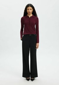 Naine seisab ettepoole, seljas burgundi punane pikakä sleeves t-särk, mustad laiad püksid, must vöö ja mustad teravatipulised kingad.