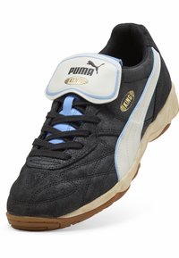 Sapatilha de camurça preta com uma faixa branca e azul clara. Possui um colarinho almofadado, atacadores pretos e uma sola de borracha. Logo da Puma exibido.