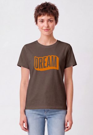 Jeune personne aux cheveux courts portant un T-shirt marron foncé avec le mot "DREAM" en grandes lettres orange, assorti à un jean bleu clair.