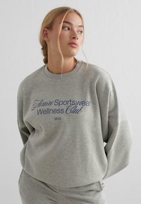 Gray cotton sweatshirt with a round neckline, dropped shoulders, and blue text reading "Flinn Sportswear Wellness Club 2013." Soft texture.