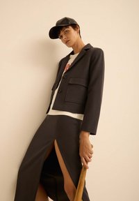 Blazer gris avec poches avant doubles, fermeture à bouton, et jupe assortie avec hautes fentes. Le modèle porte un t-shirt graphique blanc et une casquette noire.