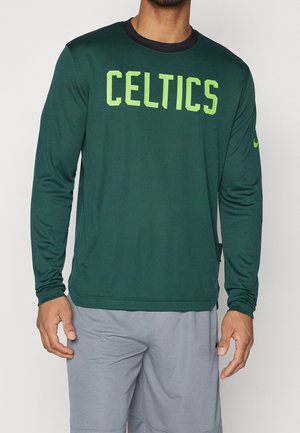 Homme portant un t-shirt vert à manches longues avec "CELTICS" en grandes lettres jaunes et un short de sport gris.