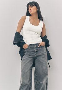 Débardeur blanc à encolure dégagée, associé à un jean gris à coupe décontractée. Le mannequin porte une veste noire posée sur un bras.