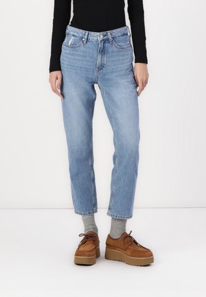 Personne portant un jean droit bleu clair, des chaussettes grises, des chaussures à plateforme marron et un haut à manches longues noir, debout devant un fond blanc.