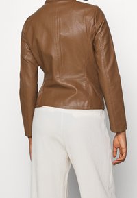 Veste en cuir brun au design ajusté, avec des coutures visibles et une texture lisse. Portée avec un pantalon large texturé de couleur claire.