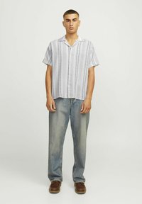Chemise légère à manches courtes avec un col, présentant des rayures verticales grises sur fond blanc. Associée à un jean délavé bleu ample et des chaussures brunes.