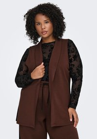 Brown, sleeveless vest in a soft material, layered over a black floral-print long-sleeve top; features a relaxed fit and open front design.