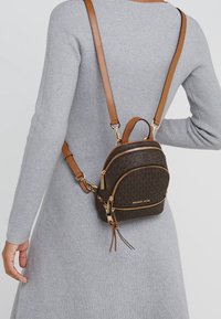 Zaino in pelle marrone con due scomparti con cerniera, dettagli in metallo dorato e spallaccio regolabile marrone, indossato a tracolla sopra un maglione grigio.