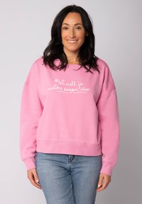 Roze sweatshirt gemaakt van zachte stof met een ronde hals, verlaagde schouders en witte tekst op de voorkant. Gecombineerd met blauwe spijkerjeans.