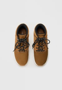 Zapatillas de gamuza marrón con cordones de patrón negro y blanco, cuello acolchado en el tobillo y suelas de goma blancas. Forro de tela transpirable.