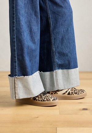 Brede blauwe jeans met rafelende zomen, gecombineerd met beige sneakers met luipaardprint op een houten vloer.