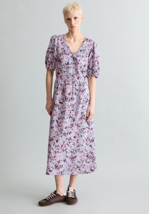 Junge Person mit kurzen blonden Haaren, die ein hellviolettes, geblümtes Midikleid und braune Sneakers vor einem schlichten Hintergrund trägt.