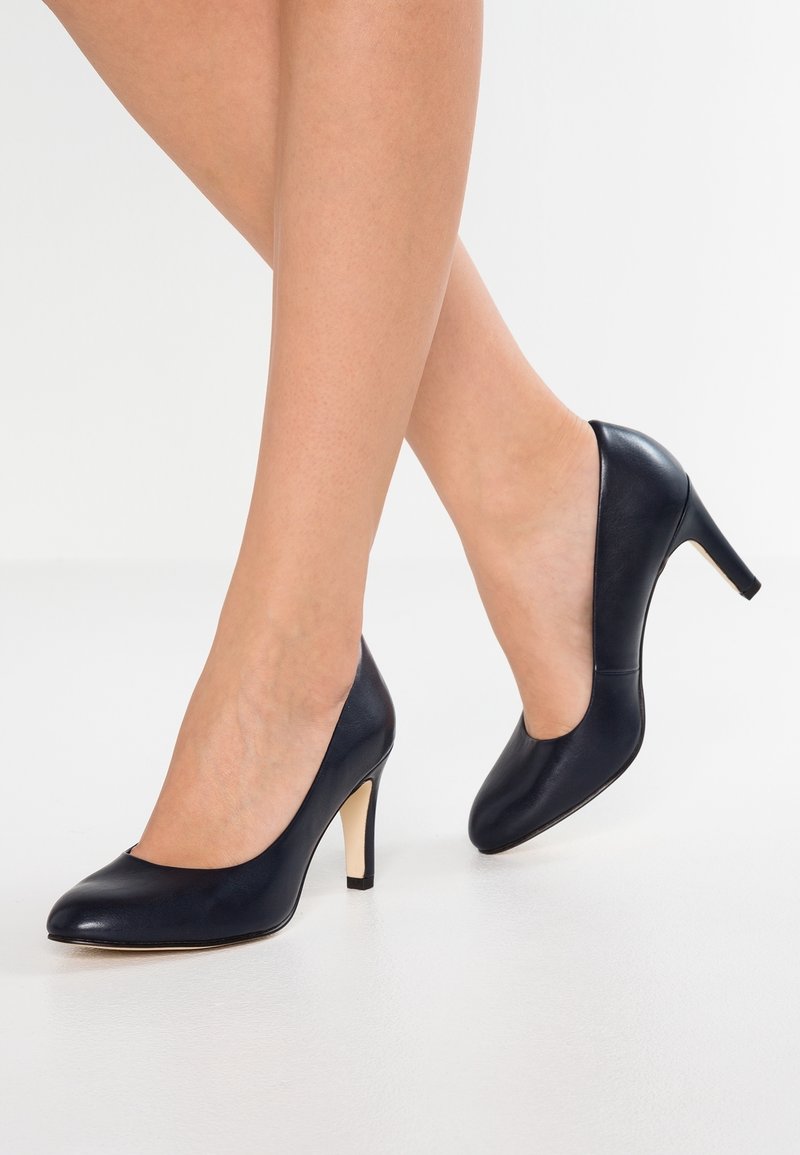 Jambes d'une femme portant des escarpins noirs en cuir à bout fermé avec des talons fins, debout sur une surface blanche lisse.