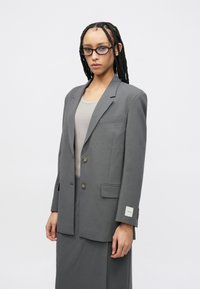 Γκρι φαρδύ σακάκι με δύο καφέ κουμπιά, πλαϊνές τσέπες και ετικέτα στο μανίκι. Το μοντέλο φοράει εφαρμοστό γκρι τοπ από μέσα.