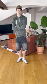 Sivana kapuca, temno modri kratki hlače z raztrganimi robovi in srebrni superge. Zaprti prostor z rožnatimi lončnicami in rjavim kavčem.