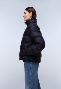 Giacca nera imbottita con colletto alto, chiusura con zip e tasche laterali; presenta una texture liscia e un design a righe orizzontali.