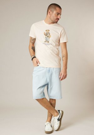 Homme tatoué portant un t-shirt beige clair avec un graphique d'ours, un short en denim clair, des baskets blanches et des bracelets, debout avec une main dans la poche.