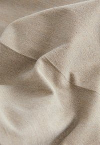 Beiger Stoff mit einer weichen Textur, der subtile Farbvariationen und Falten zeigt. Das Material wirkt leicht und gewebt.