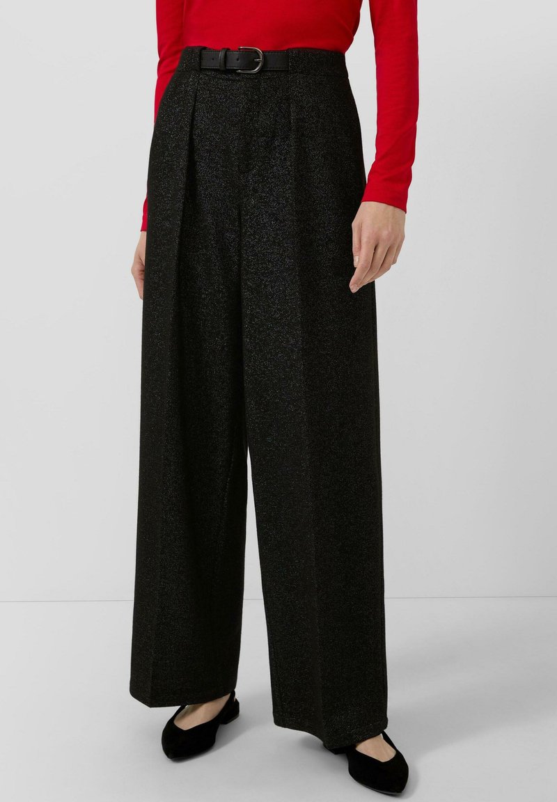 Zwarte wijde broek met een schitterende textuur, voorzien van een aansluitende taille en een zwarte riem. Gepaard met een rode top met lange mouwen en zwarte flats.
