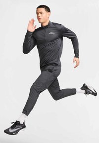 Dunkelgraues Laufoutfit mit einem Reißverschluss-Top und schmal zulaufender Hose, gefertigt aus flexiblem Stoff. Schwarze Laufschuhe mit weißen Akzenten.