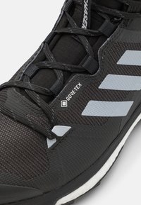 Zapato de senderismo negro con un upper texturizado, múltiples lazos de cordones y tres rayas azul claro. Incluye una etiqueta visible de GORE-TEX.
