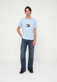 Ανοιχτό μπλε βαμβακερό t-shirt με το λογότυπο "Tommy Hilfiger" και γραφικό σημαίας στο μπροστινό μέρος, συνδυασμένο με σκούρους τζιν και μαύρα παπούτσια.