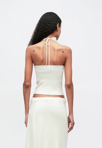 Kvinne med langt krøllete hår som har på seg en hvit halterneck topp og skjørt, som viser ryggen sin med to små firkantede tatoveringer på skuldrene.