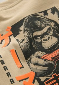 Beige t-shirt featuring a black gorilla graphic wearing headphones, holding a game controller, with bright orange text accents.