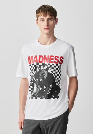 Junger Mann trägt ein weißes T-Shirt mit rotem "MADNESS"-Schriftzug und einem schwarz-weißen Foto von fünf Männern in Anzügen vor einem karierten Hintergrund.