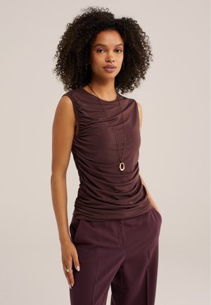 Femme aux cheveux bouclés portant un haut sans manches froncé violet foncé, un pantalon assorti et un long collier avec un pendentif en or.