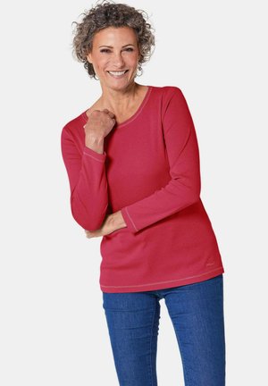 Chemise rouge à manches longues en tissu côtelé avec des coutures contrastées et un col en scoop, associée à un jean bleu. Le mannequin pose avec un sourire.