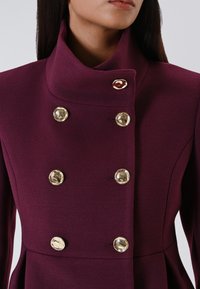 Cappotto doppiopetto in un profondo bordeaux. Realizzato in tessuto testurizzato, presenta un colletto alto e bottoni rotondi tono oro per la chiusura.
