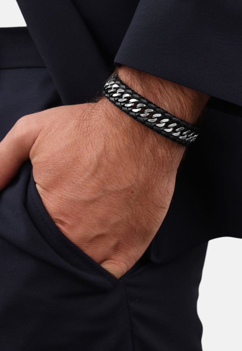Zwarte gevlochten leren armband met zilveren metalen schakels; gedragen om een pols. De achtergrond toont een donker colbert met een zichtbare zak.