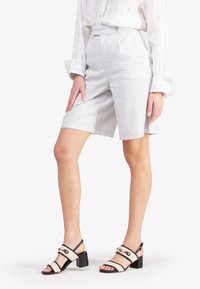 Pantaloni corti grigio chiaro su misura con un tessuto testurizzato e una vestibilità ampia, abbinati a sandali neri e bianchi con tacco e dettagli dorati.