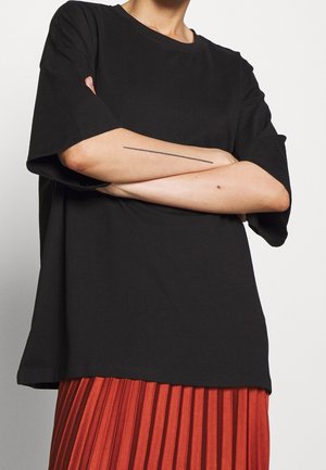 Osoba nosząca czarną oversize koszulę z krótkim rękawem i czerwoną plisowaną spódnicę, ze skrzyżowanymi ramionami, pokazuje cienki, liniowy tatuaż na lewym przedramieniu.