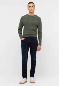 Zielona prążkowana koszula henley z długim rękawem, z trzema guzikami i wyszywanym logo, w połączeniu z ciemnoniebieskimi wizytowymi dżinsami i beżowymi trampkami.