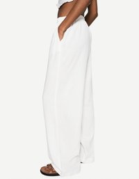 Une personne se tient habillée de pantalons blancs à jambes larges et d'un haut court, les mains dans les poches, assortis avec des sandales marron.