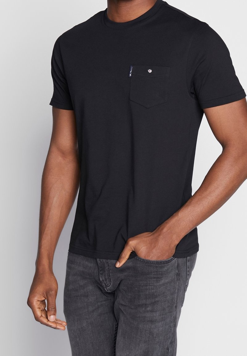 Camiseta negra de manga corta hecha de algodón, con un bolsillo en el pecho y detalle de botón. Diseño ajustado con escote redondeado.