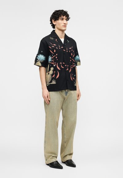 Άνδρας που στέκεται, φορώντας μαύρο κοντομάνικο πουκάμισο με σχέδιο κάκτων και λουλουδιών, άνετο μπεζ παντελόνι και μαύρα μυτερά παπούτσια.