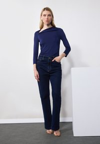 Mørkeblå langermet topp med drapert utringning, kombinert med høyt liv mørke denimmansje. Modellen står med hånden i lommen og har på seg gullsandaler.